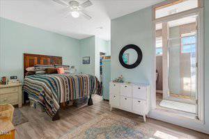 A bedroom with a wooden bed featuring a patterned comforter, a ceiling fan, a white cabinet with drawers, a round mirror on the wall, and an en suite bathroom with a glass shower door.