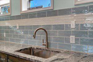 A kitchen sink with a silver faucet set in a stone countertop, blue and white subway tile backsplash, and electrical outlets on the wall. A sign in the window reads "BE AWESOME TODAY.