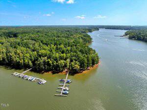 Aerial view of a lake bordered by dense green forest, featuring several docks with boats moored along the shoreline and a few boats visible on the water under a clear blue sky.