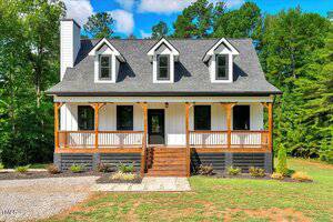 This charming two-story house at 692 Port Drive, Henderson features three dormer windows, a front porch with wooden railings and columns, a central staircase, and is surrounded by green trees and a gravel walkway.