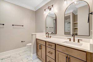 A modern bathroom with a double vanity, two mirrors, brown cabinets, black fixtures, a toilet, and marble tile flooring. The walls are beige, and there are three light fixtures above the mirrors.