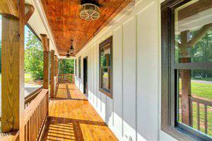 A wooden porch with a polished floor, white board-and-batten siding, brown railing, and black-trimmed windows, viewed in daylight with greenery and a road visible in the background.