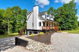 A modern two-story white house with black trim and a covered front porch sits on a gravel driveway, surrounded by landscaped rocks and mulch, with trees in the background under a blue sky.