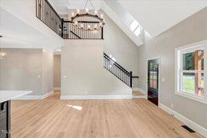 A spacious entryway and living area with light wood floors, beige walls, a black metal staircase railing, large windows, and a modern circular chandelier hanging from a high ceiling.