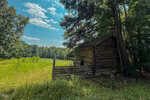 A wooden log cabin stands on the edge of a grassy field on 24 Acres Bob Daniel Road, partially shaded by tall trees. The sky is mostly clear with some scattered clouds, and dense forest is visible in the background near Oxford.