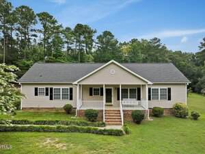 Charming single-story beige home at 2138 Old Mill Farm Road, Franklinton, with white trim, a covered front porch, black shutters, and brick steps. Surrounded by green lawn, bushes, and tall trees in the background.