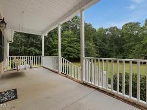 A covered front porch with white railings, a porch swing on the left, and concrete flooring overlooks a grassy yard bordered by trees under a partly cloudy sky.