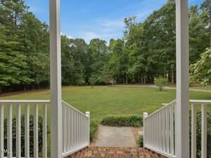 View from a porch with white railings and brick steps leading down to a concrete path and a large grassy yard, bordered by tall trees under a blue sky.