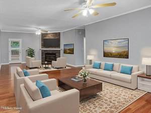 A spacious living room with light beige sofas, blue accent pillows, a patterned rug, wood flooring, two ceiling fans, a fireplace with a TV above it, and two landscape paintings on gray walls. A door leads outside.