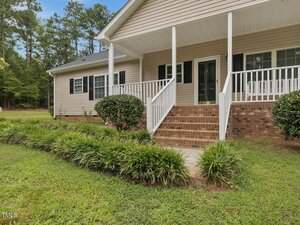 A beige single-story house with white trim, a covered front porch, brick steps, white railings, and black shutters. The yard has bushes, green grass, and a row of plants along the walkway. Trees are in the background.