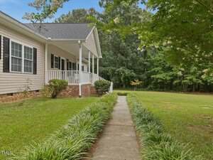 A concrete walkway bordered by green plants leads to a beige single-story house with white trim, a brick foundation, black shutters, and a covered front porch. The yard is grassy with trees in the background.