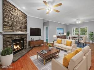 A staged living room with a stone fireplace, wall-mounted TV, two beige sofas with yellow cushions, and a wooden coffee table. In the background, a dining area with a table, chairs, and large window is visible.