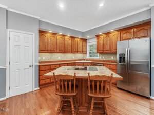 A kitchen with wooden cabinets, stainless steel refrigerator, a central island with a built-in cooktop, two wooden chairs, hardwood floors, and a window above the sink.