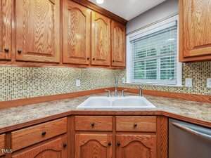 A kitchen with wood cabinets, mosaic tile backsplash, beige countertop, white double sink, and a window with blinds above the sink. The lower right corner shows part of a stainless steel dishwasher.