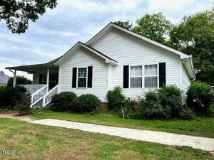 Charming single-story white house with black shutters at 15296 N C 39 Highway, Henderson. Features a covered front porch, steps to the entrance, manicured lawn, shrubs, sidewalk in front, and mature trees surrounding the home.