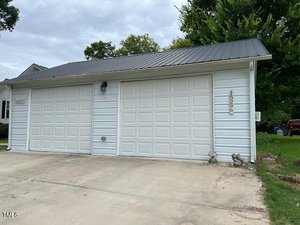 A detached two-car garage with white siding, two white garage doors, a black metal roof, and house number 1526. The driveway is concrete, and there is greenery and trees in the background.