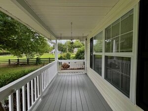 A covered front porch with gray flooring, white railings, and large windows on the right. A ceiling fan and hanging light fixture are overhead. Trees, a wooden fence, and grass are visible in the background.