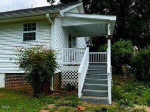 A white house with a covered porch and gray steps leading up to the front door, surrounded by overgrown grass and bushes. A small table and chairs are visible in the yard to the right.