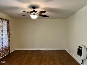 An empty room with light-colored walls, hardwood flooring, a ceiling fan with a light fixture, patterned curtains on one window, and an electric wall heater on the right side.