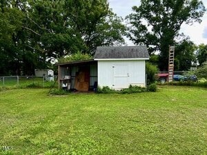 A small white shed with a gray roof stands on a grassy lawn. The shed has double doors, an open lean-to area with various items stored inside, and is surrounded by trees and a chain-link fence. A ladder leans on the right side.