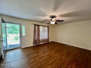 Empty living room with wood flooring, a ceiling fan with lights, a large window with patterned curtains, and an open door leading outside; walls are painted a light color.