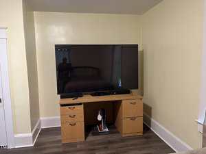 A flat-screen TV is placed on a wooden desk with drawers in a corner of a room with light yellow walls and dark wood flooring. A game console and a remote are on the desk, and cords are visible underneath.