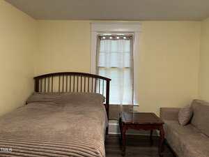 A bedroom with light yellow walls features a bed with a wooden headboard and beige bedding, a wooden side table, a beige couch, and a window with a curtain letting in natural light.