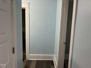 A small hallway with light blue walls, white trim, and dark wood flooring. There are three doorways visible, one partially open on the right and another leading to a darker room on the left.