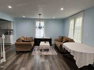 A living room with two brown sofas, a small coffee table with a lace cloth, a dark wood sideboard, and a round dining table covered with a white tablecloth. The room has blue walls, wood flooring, and two large windows.