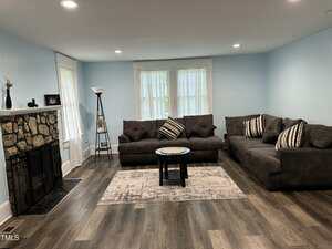 A living room with light blue walls, hardwood floors, a stone fireplace, two dark gray sofas with striped pillows, a small round coffee table, a rug, and a floor lamp by a window with sheer curtains.