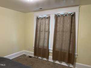 A beige-carpeted room with light yellow walls, two brown curtains covering a double window, white trim, a vent in the ceiling, and visible electrical cords on the floor next to the window.