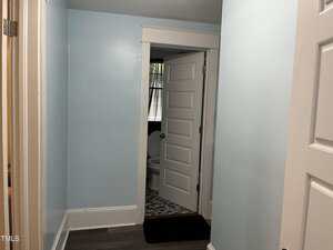 A hallway with light blue walls and dark flooring leads to a bathroom with a white door ajar. Inside the bathroom, a toilet and a window with a curtain are visible.
