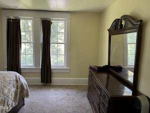 A bedroom with two large windows with brown curtains, a bed with a beige and white blanket, beige carpet, and a wooden dresser with an attached mirror. Sunlight is coming through the windows.