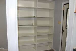 A built-in white shelving unit with eight shelves is installed against a light gray wall. An empty closet rod and some cords are visible on one shelf. A door with a window is on the right.