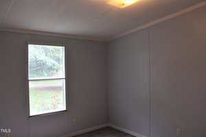 A small, empty room with light blue walls, a single window showing greenery outside, a white ceiling with a ceiling light, and gray flooring. There are two electrical outlets visible near the window.