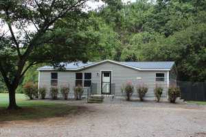 A single-story gray house with a metal roof at 350 Hamp Falkner Road, Henderson. Several windows and a small front porch with steps overlook bushes lining the front, while a gravel driveway leads to the entrance. Trees and greenery surround the property.