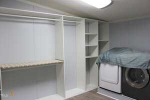 Built-in open closet with shelves and a hanging rod on the left, next to a washing machine and dryer partially covered by a quilted blue-gray cover in a laundry room with light gray walls and wood flooring.
