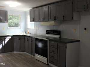 A kitchen with gray cabinets, black countertops, a white stove with a matching hood, a stainless steel sink under two windows, and light wood flooring. The walls are light blue, and there are no visible appliances on the counters.