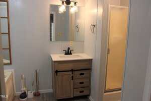 A bathroom features a vanity with a rectangular mirror, black faucet, and wooden cabinet, next to a shower with a frosted glass door. Cleaning tools, including a plunger and toilet brush, are on the left.