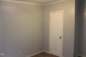 A plain room with light gray walls, a white door with a silver handle, light-colored wood flooring, and a single electrical outlet on the left wall. The room appears empty and undecorated.