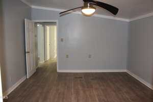 Empty room with gray walls, wood-look flooring, a ceiling fan with a light, and an open door leading to a hallway with additional doors visible.