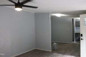 A room with light gray walls, a dark ceiling fan, and wood-style flooring. An adjacent area contains a paint bucket and supplies, and part of a kitchen is visible in the background.