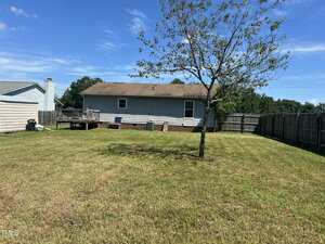 A backyard with a mowed lawn, a single tree, wooden fencing, and a light blue house with a deck and visible foundation. A clear blue sky is overhead.
