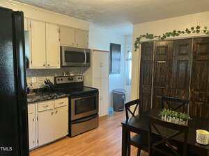 A kitchen with white cabinets, stainless steel appliances, a black refrigerator, a dining table with four chairs, and dark wooden closet doors. Decorative greenery hangs above the closet. The floor is light wood.