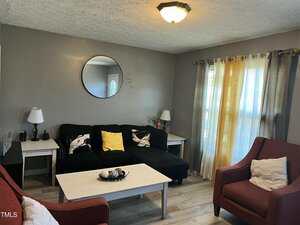 A small living room with gray walls, a black sectional sofa with patterned and yellow pillows, two red armchairs, a white coffee table, a round mirror on the wall, and sheer yellow curtains on the window.