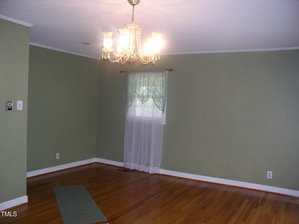 An empty room with green walls, a wooden floor, a window with sheer curtains, and a ceiling chandelier light fixture. A small floor mat is near one wall.