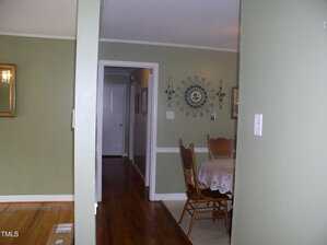 A dining area with a round table covered by a lace tablecloth and four wooden chairs is visible. A wall clock and candles are mounted on the wall. A hallway and several doors are seen in the background.
