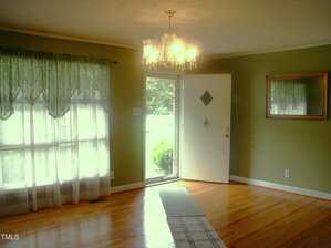 A room with hardwood floors, green walls, a window with sheer curtains, a wall mirror, and an open white door letting in natural light. A ceiling chandelier is turned on above a runner rug.