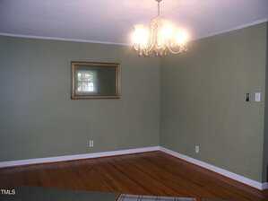 A mostly empty room with green walls, a wooden floor, a ceiling-mounted chandelier light, and a framed mirror on one wall. There is a light switch and an electrical outlet visible on the right wall.