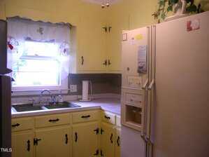 A small kitchen with yellow cabinets, a double sink under a window with decorative curtains, a beige refrigerator with notes and magnets, and a white kettle on the countertop. There are plants above the fridge.